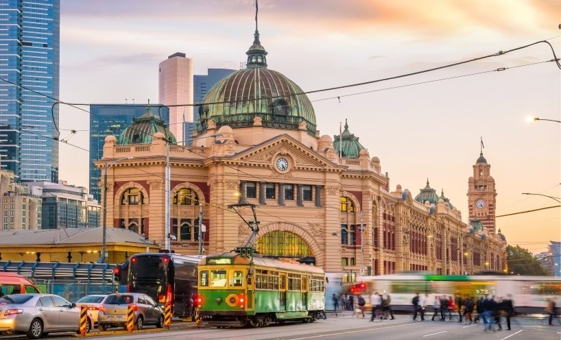 Trams and pedestrians crossing the intersection in front of Flinders St Station at dusk.