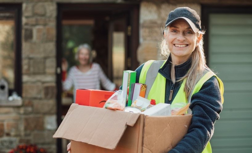 Smiling woman in yellow hi-viz vest carrying groceries to another woman.